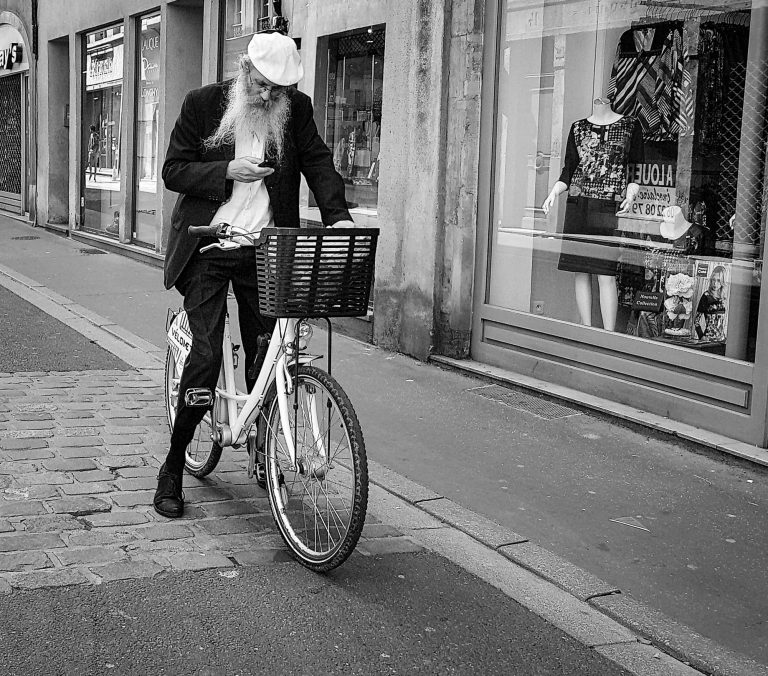 Black and white photo of a man pushing a bicycle with a basket along a cobbled street