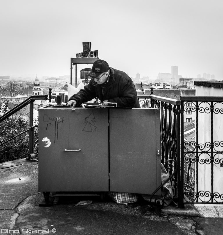 Black and white street photo of a man leaning over a metal container by a railing