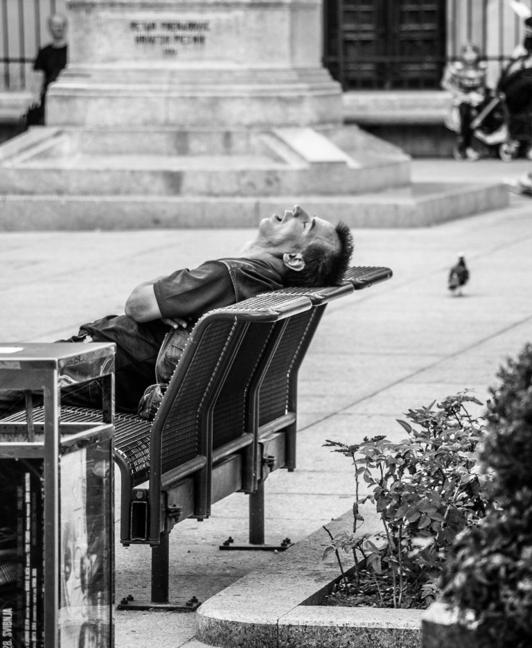 Black and white street photo of a man lying asleep on a bench