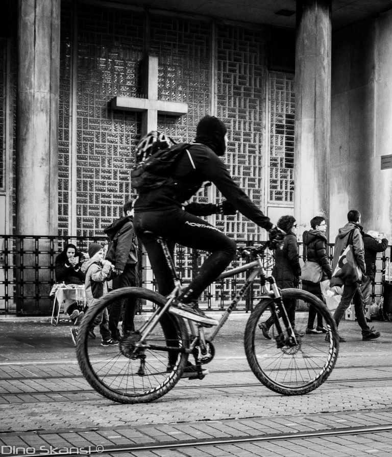 Black and white street photo of a cyclist riding past a building with a cross