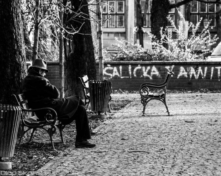 Black and white photo of a man in a hat sitting on a bench in a cobbled square
