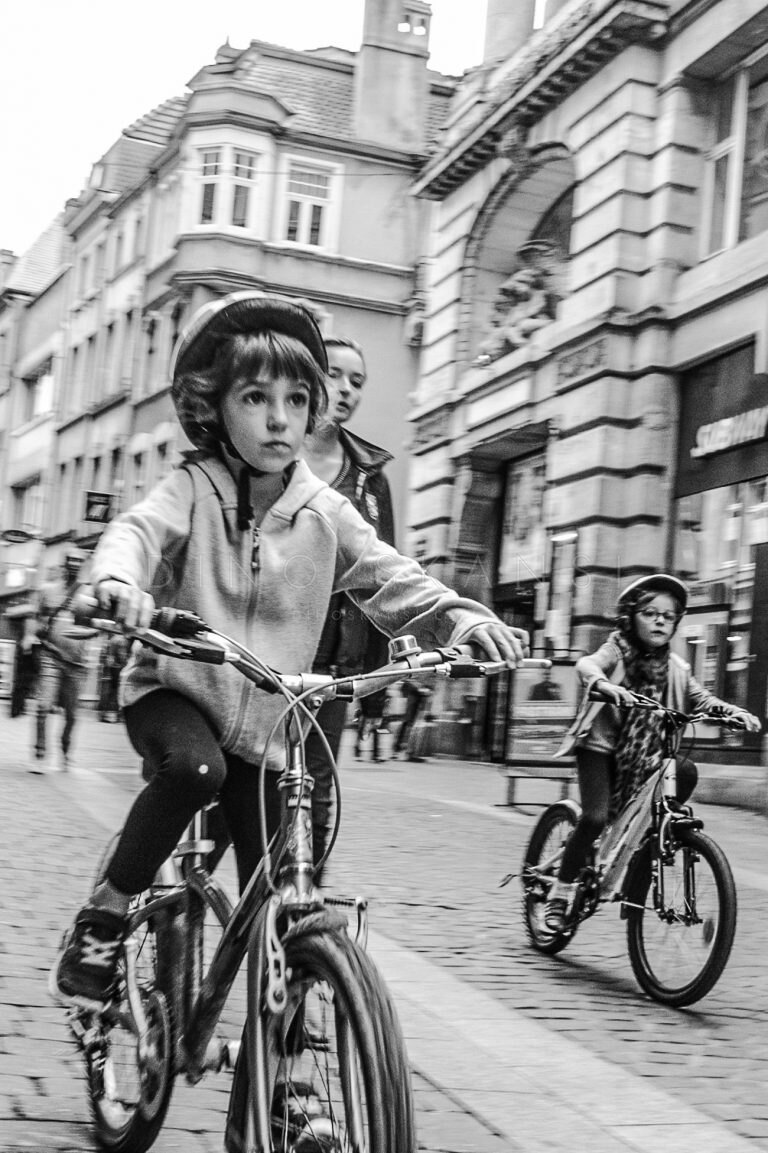 Black and white street photo of two children in helmets riding bicycles