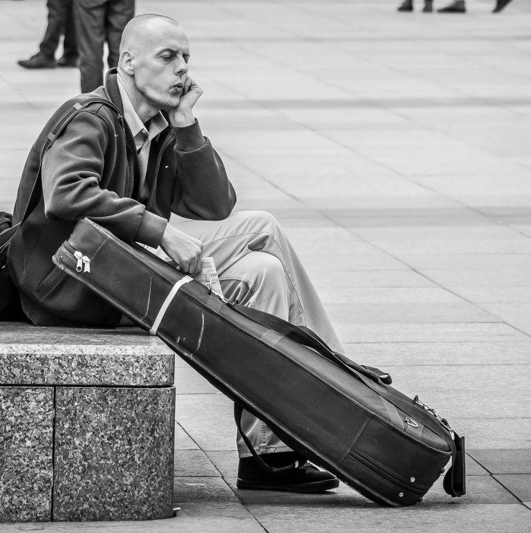 Black and white street photo of a man sitting on a stone block beside a guitar case