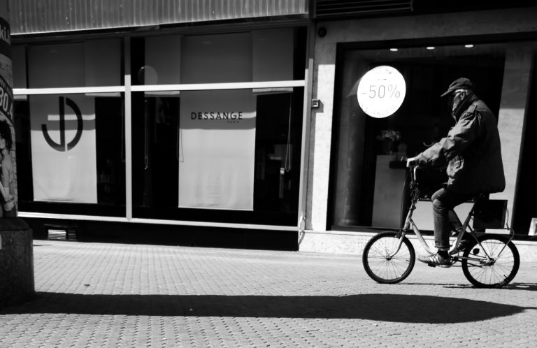Black and white street photo of a cyclist passing a shop window with a sale sign