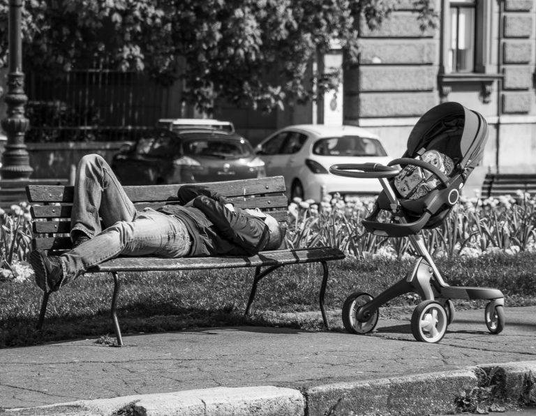Black and white street photo of a man lying on a park bench beside a pushchair