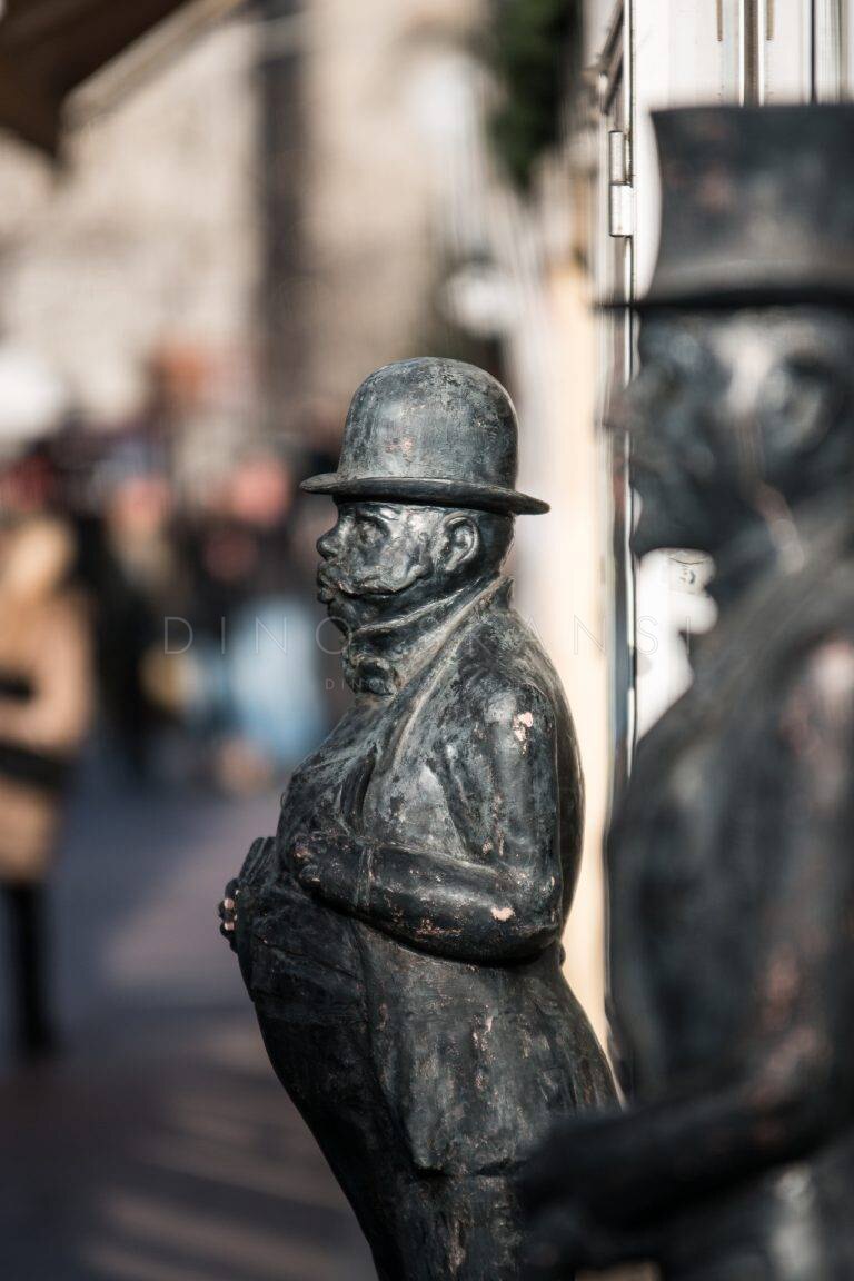 Colour photo of a bronze statue of a man in a bowler hat on a busy street