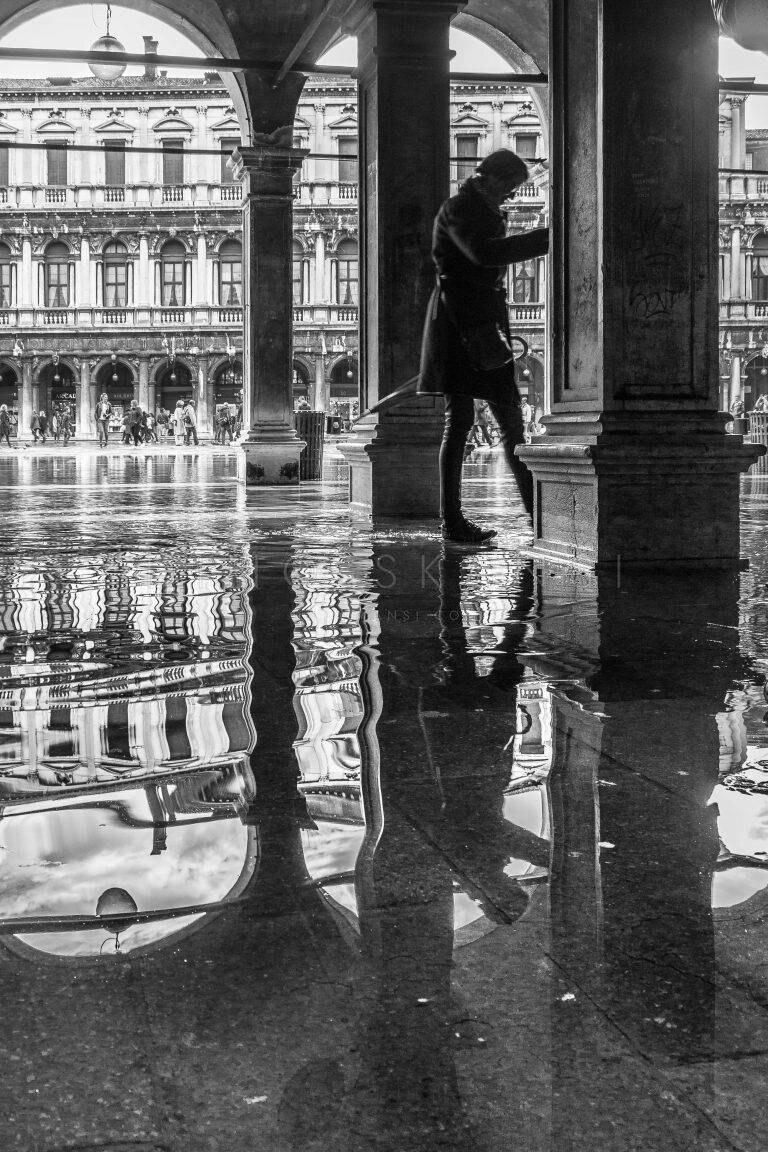 Black and white photo of a figure walking through a flooded arcade in Venice