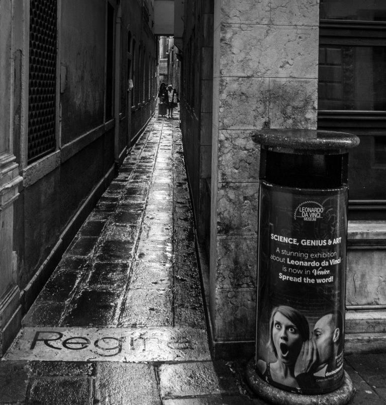 Black and white photo of a narrow Venetian alley with a litter bin in the foreground