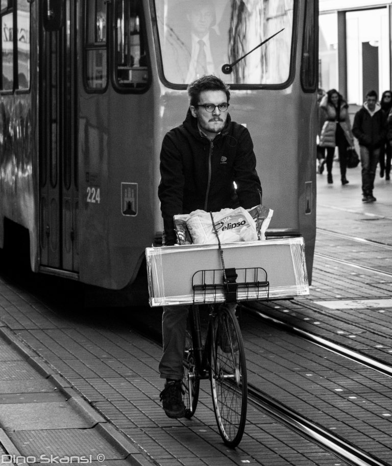 Black and white street photo of a man cycling beside a tram with a box on his bicycle