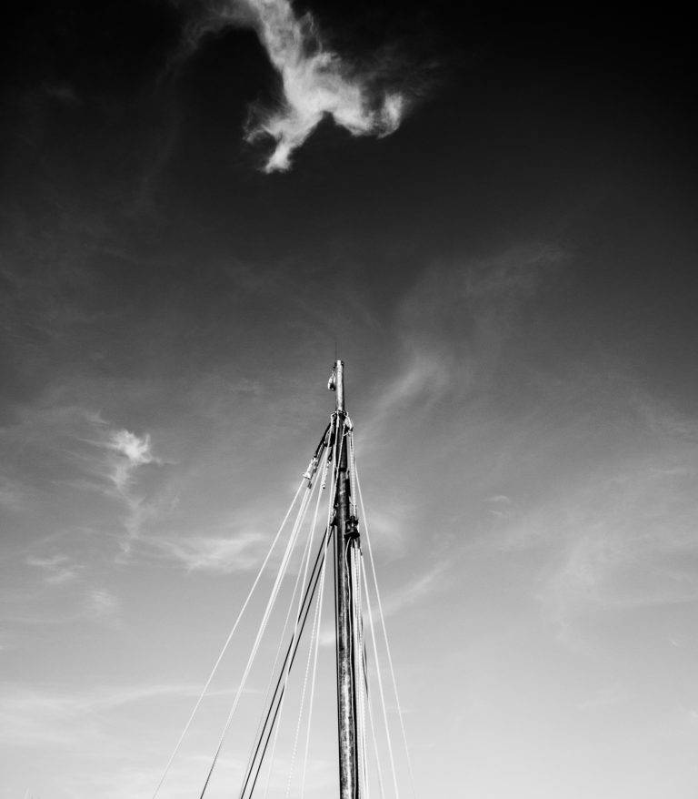 Black and white photo of a sailing boat mast against a cloudy sky