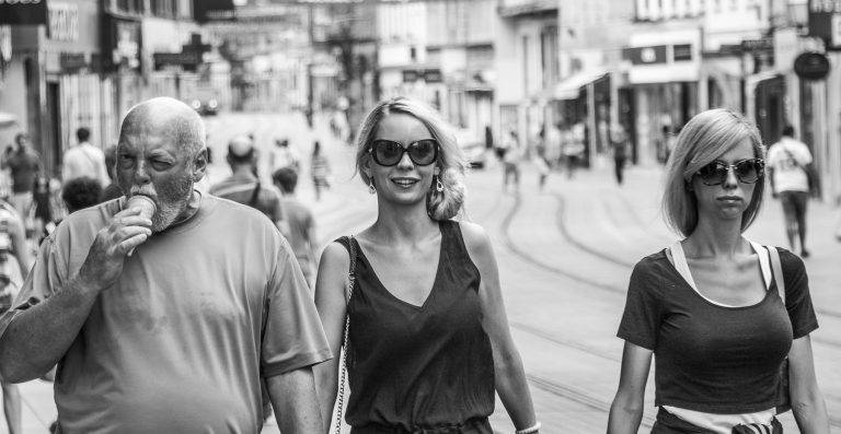 Black and white street photo of a smiling woman in sunglasses walking along a busy street