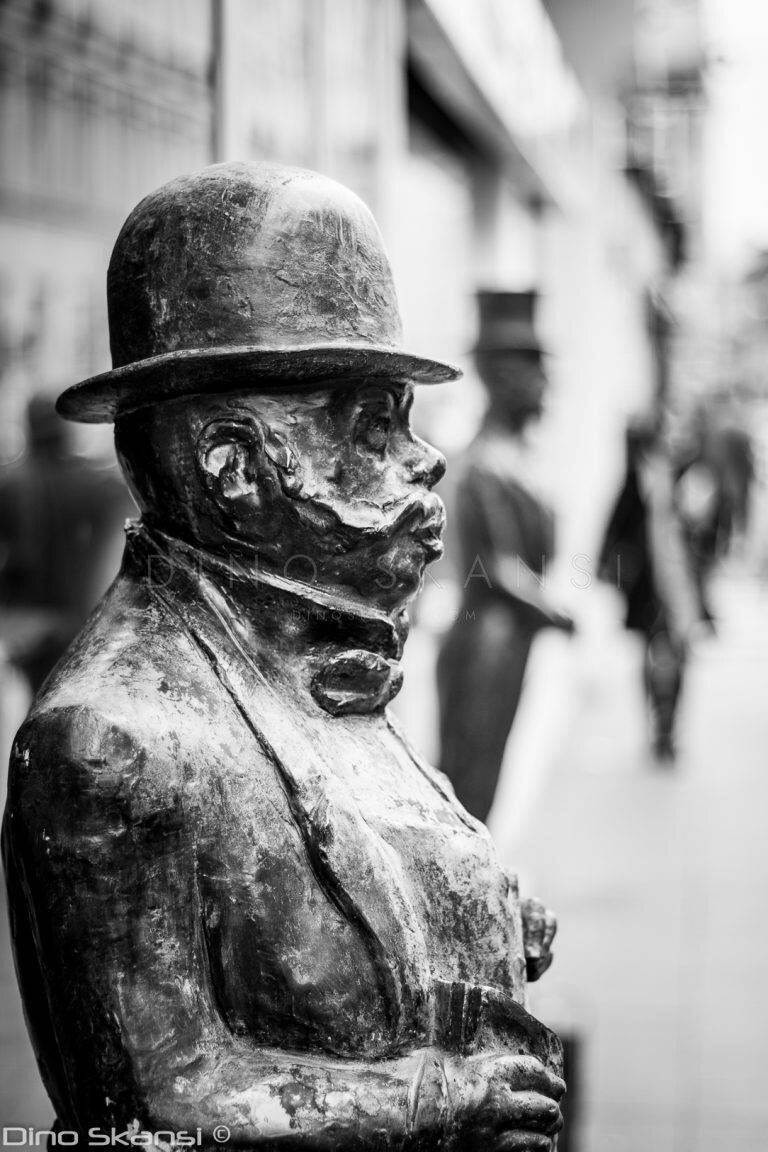 Black and white close-up of a bronze statue of a man in a bowler hat on a busy street