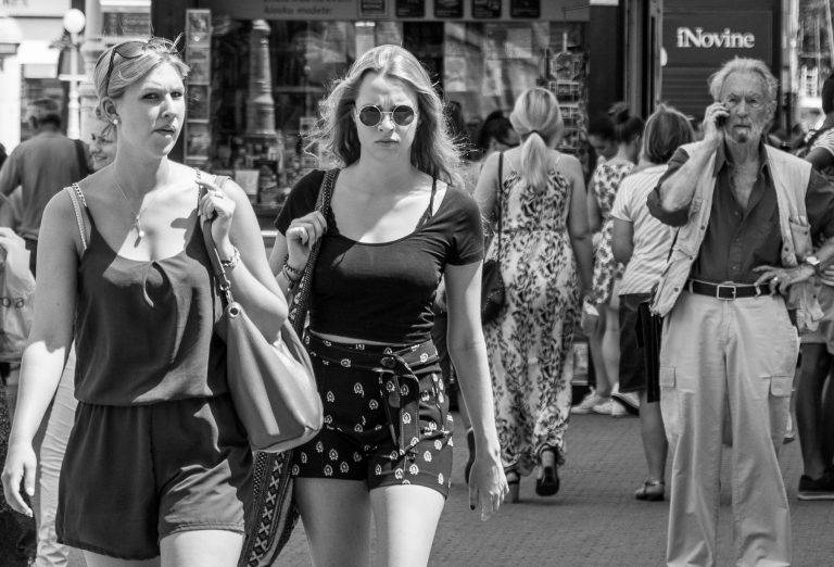 Black and white street photo of a woman in sunglasses and shorts walking through a crowd