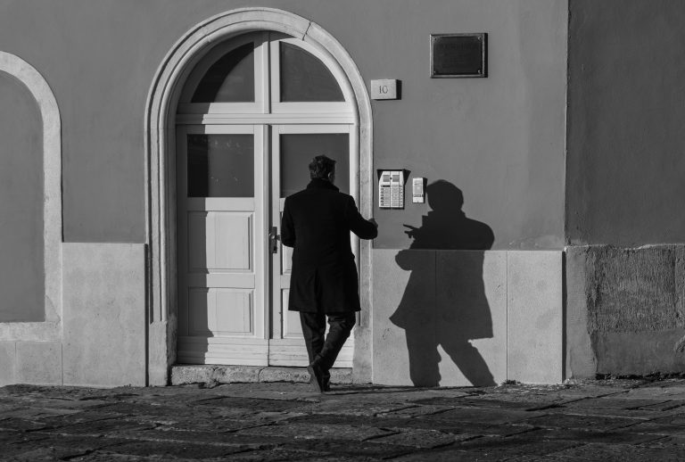 Black and white photo of a man entering an arched doorway beside his shadow on the wall