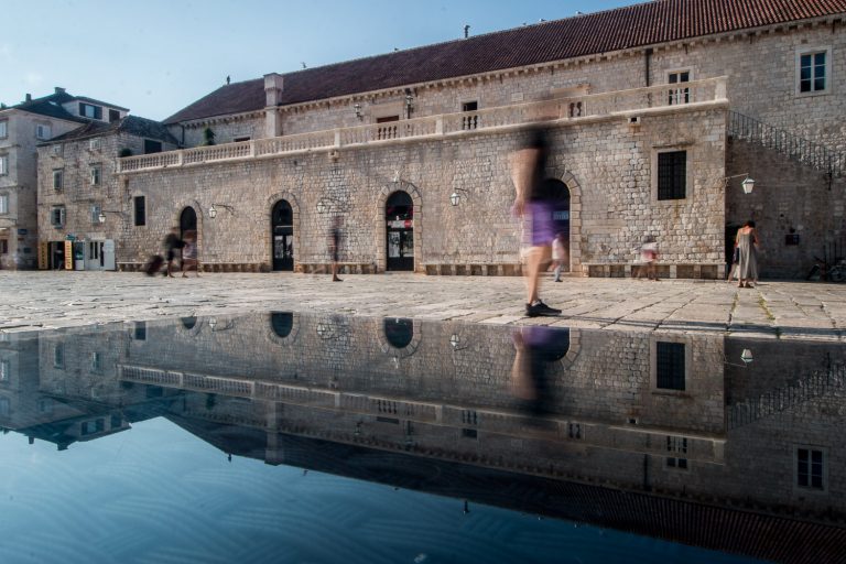 Colour street photo of a stone building reflected in a wet square, with a blurred passer-by