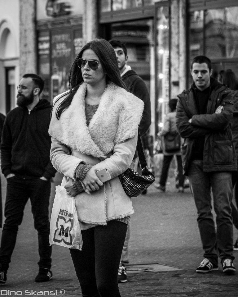 Black and white street photo of a woman in a light coat and sunglasses walking through a crowd