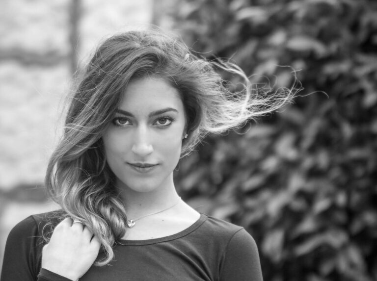 Black and white portrait of a woman with hair blowing in the wind beside a hedge