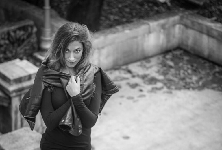 Black and white portrait of a woman holding her leather jacket closed, standing by a stone wall