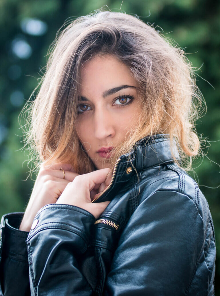 Colour portrait of a woman in a leather jacket with her hand at her chin, green background
