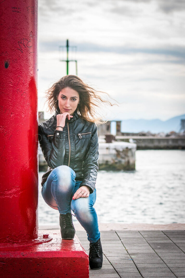 Colour portrait of a woman in a leather jacket and jeans leaning on a red harbour post