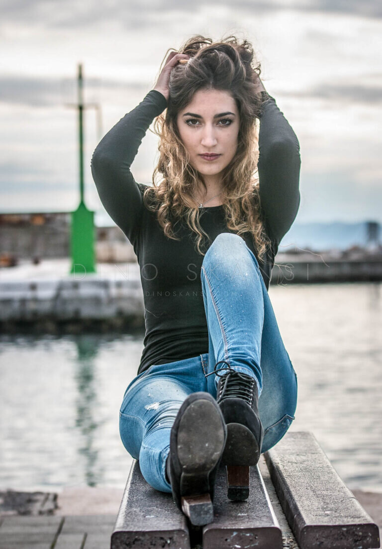 Colour portrait of a woman sitting on a harbour wall with her arms raised